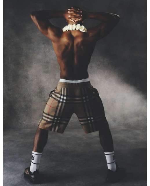 Man wearing Burberry plaid shorts and white socks poses with hands behind head. He has dark skin, muscular build, and white floral lei.