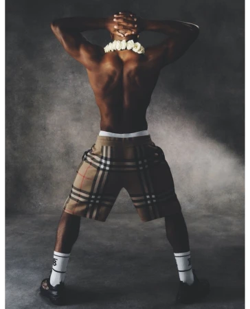Man wearing Burberry plaid shorts and white socks poses with hands behind head. He has dark skin, muscular build, and white floral lei.