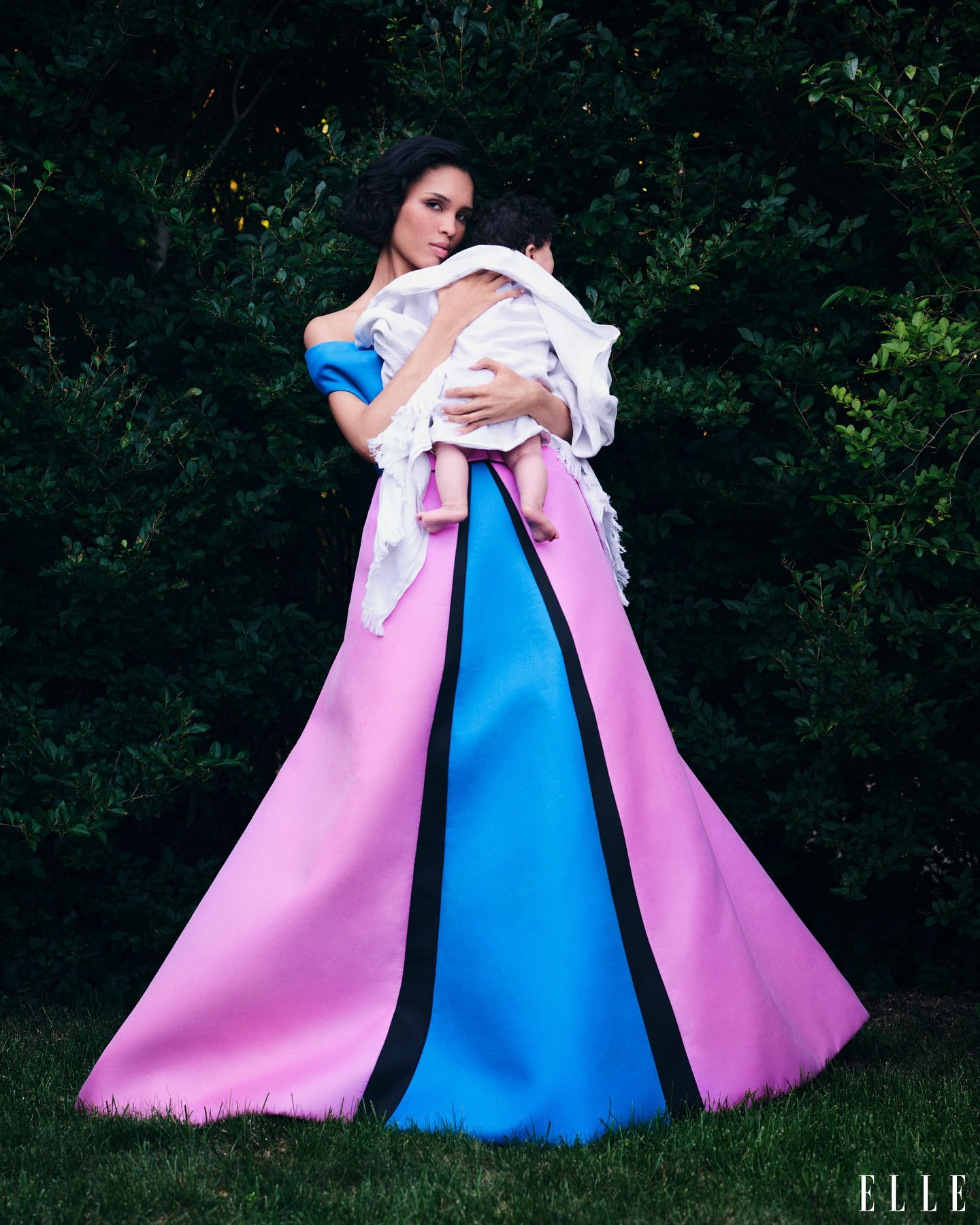 Woman in a pink and blue gown holds a baby in a lush green outdoor setting. The woman has dark hair and the baby is wrapped in white.