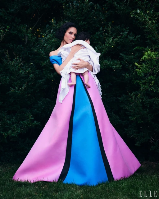 Woman in a pink and blue gown holds a baby in a lush green outdoor setting. The woman has dark hair and the baby is wrapped in white.