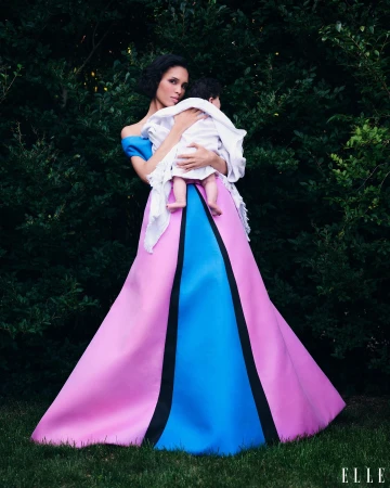 Woman with dark hair holding a baby while wearing a vibrant pink, blue, and black ballgown outdoors against dark foliage.