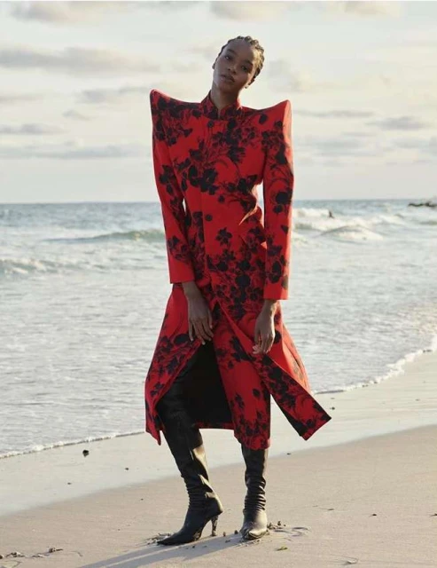 Woman on beach in striking red floral-print coat and black heeled boots poses against the ocean backdrop under an overcast sky.