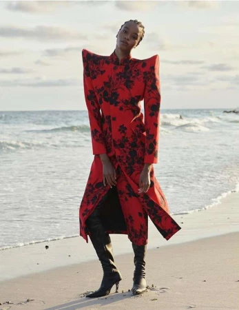 Woman on beach in striking red floral-print coat and black heeled boots poses against the ocean backdrop under an overcast sky.