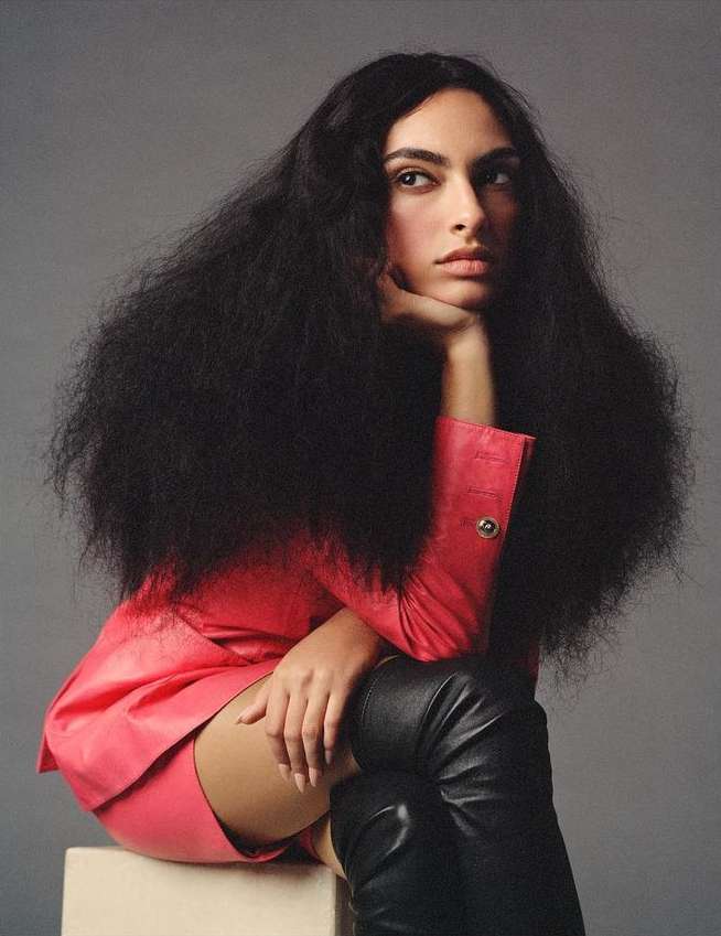 Woman with long black hair posing in a pink suit and black boots sitting on a cube in a photo studio against a grey background.
