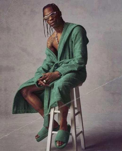 A man wearing a robe and sunglasses sits on a white stool in a photo studio against a grey backdrop, looking at the camera.