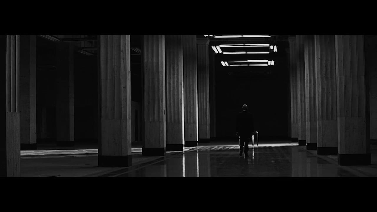 A lone figure walks down a hall lined with columns in a monochromatic color palette, illuminated by light from overhead fixtures.