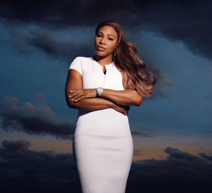 Portrait of Serena Williams in a white dress with arms crossed, posing against a cloudy sky background, wearing a wristwatch.