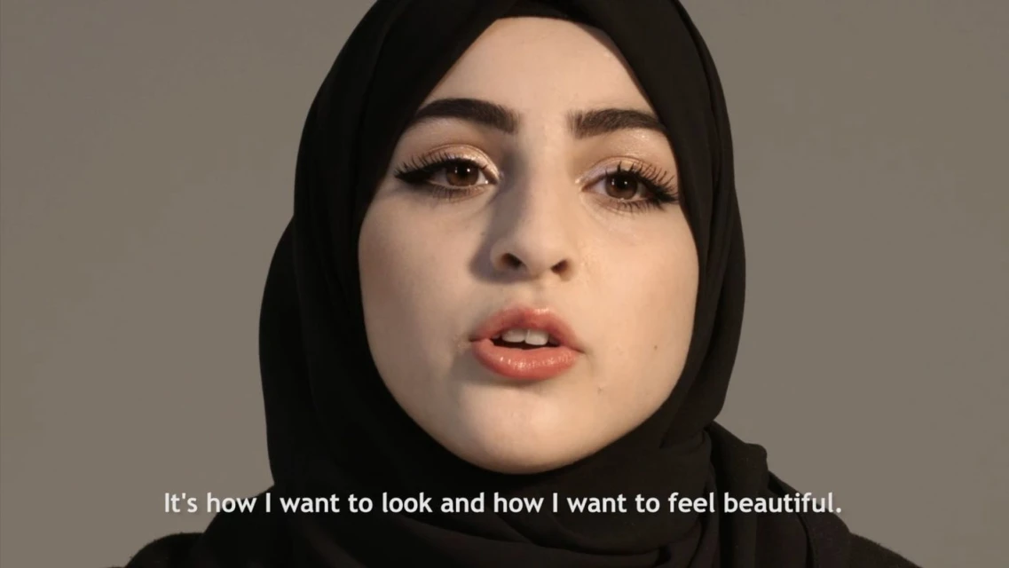 Close-up portrait of a young woman with brown eyes and dark hair wearing a black hijab against a plain grey background.