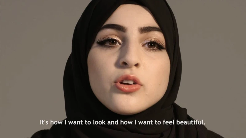 Close-up portrait of a young woman with brown eyes and dark hair wearing a black hijab against a plain grey background.