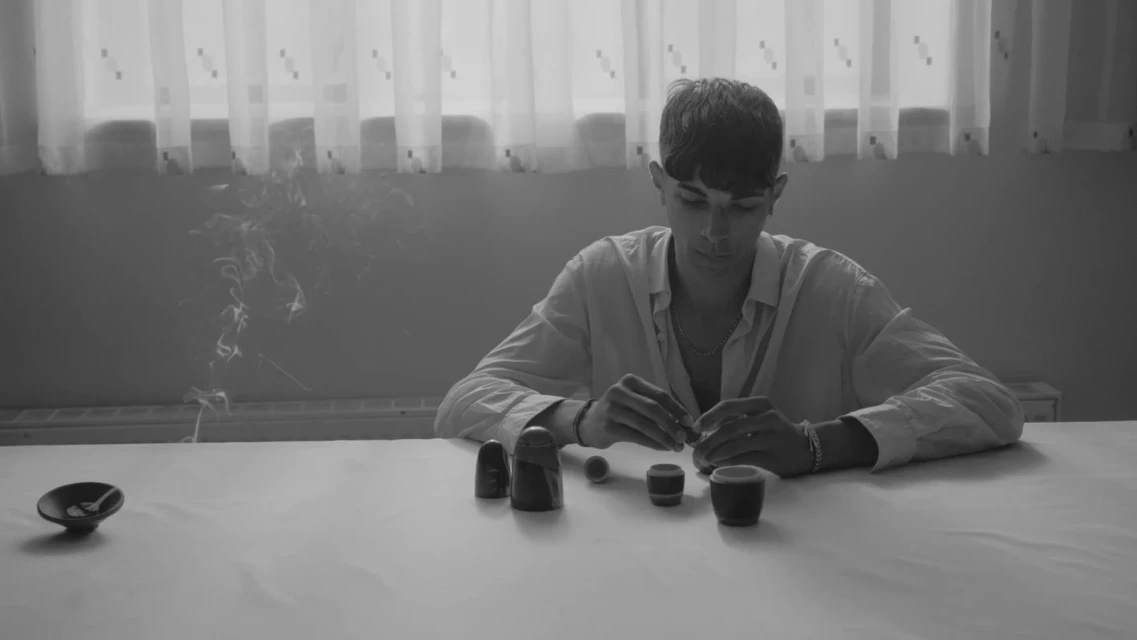 Monochrome image of a young man sitting at a table with Russian nesting dolls, smoke rising in background, creating a contemplative indoor scene.