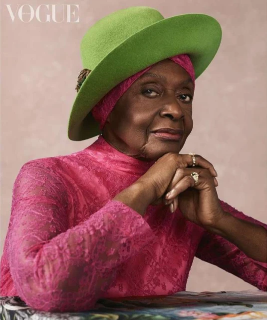 Elegant older woman with dark skin wearing a lime green hat and pink lace dress. A high-fashion editorial style portrait. Vogue logo is present.