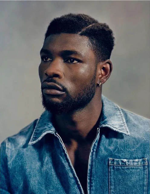 Portrait of a stylish Black man with short textured hair, wearing a denim shirt in a studio setting. Head and shoulders.