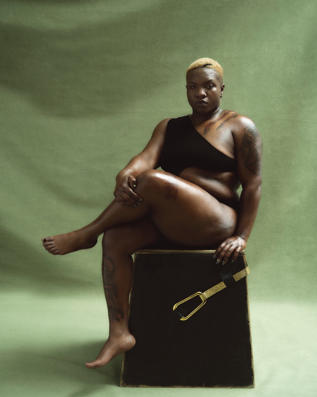 Portrait of a black woman with a blonde buzz cut, wearing black clothing, sitting on a box, set against a green background.