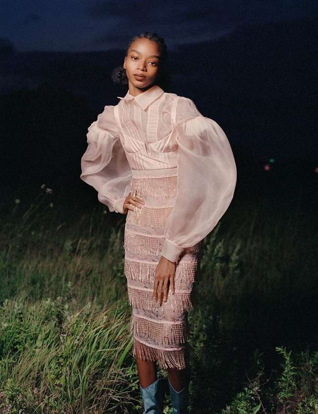 A woman poses in a sheer, blush-toned dress with a fringe skirt, paired with turquoise boots, set in a grassy field under a night sky.