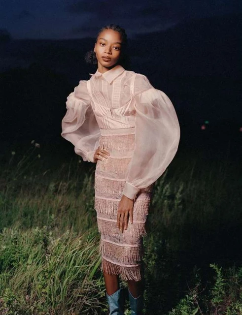 A woman poses in a sheer, blush-toned dress with a fringe skirt, paired with turquoise boots, set in a grassy field under a night sky.
