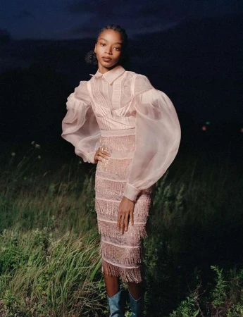 A woman poses in a sheer, blush-toned dress with a fringe skirt, paired with turquoise boots, set in a grassy field under a night sky.