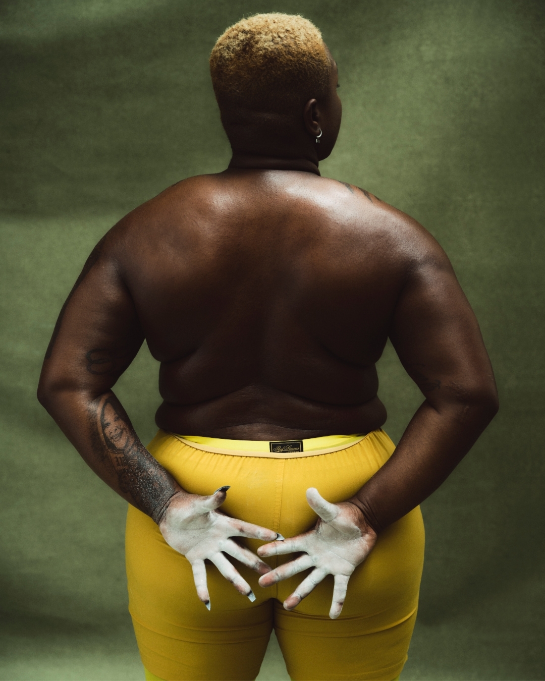 Back view of African-American adult female with blonde hair and yellow trousers against a green backdrop, showing partial vitiligo.