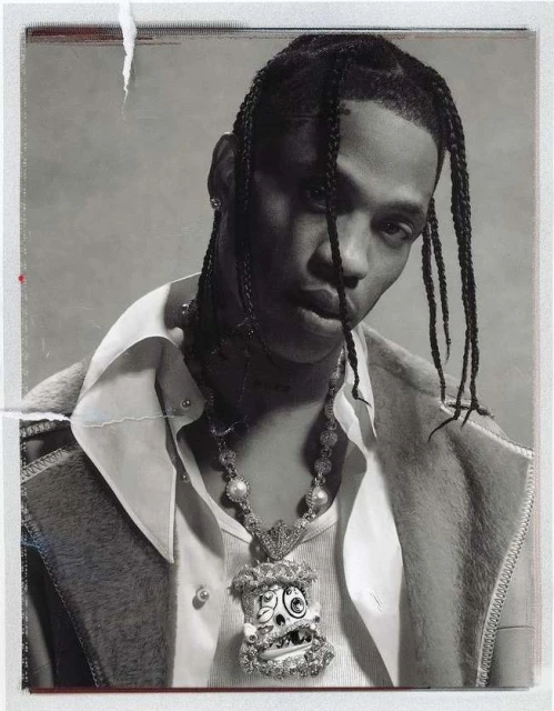Portrait of a Black male with braided hair wearing a white shirt and a large pendant necklace against a light gray backdrop.