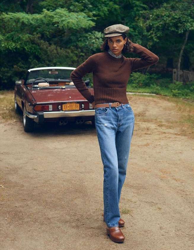 Adult woman in a brown sweater and jeans standing next to a red convertible on a dirt road with a forest background.