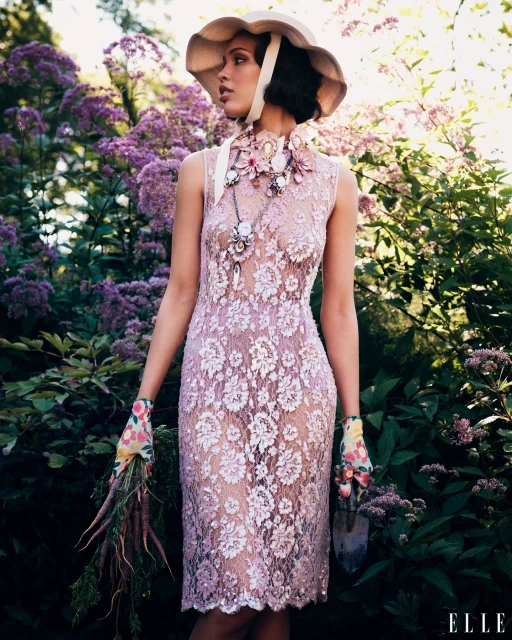 Woman in lace dress and straw hat stands among purple flowers, holding carrots and trowel in a garden for fashion editorial.