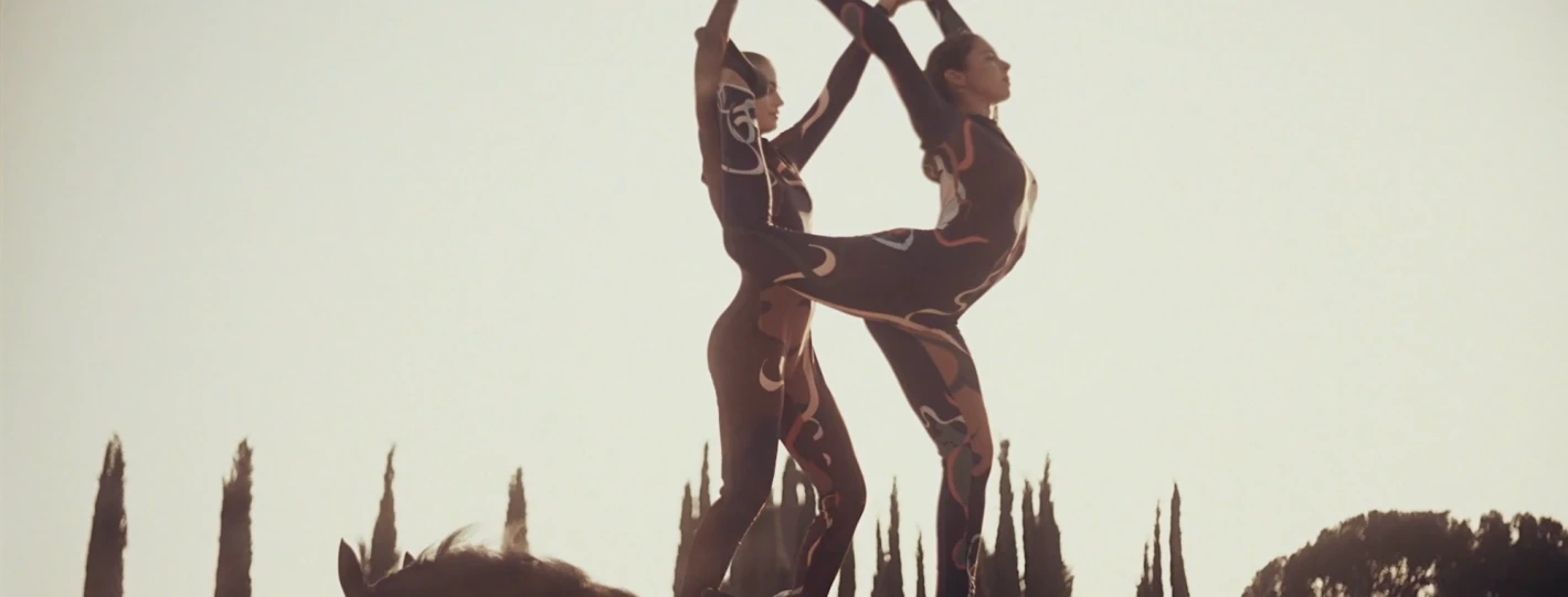 Two women in patterned bodysuits perform an acrobatic move on a horse against a bright, sunny sky in a rural setting with trees.