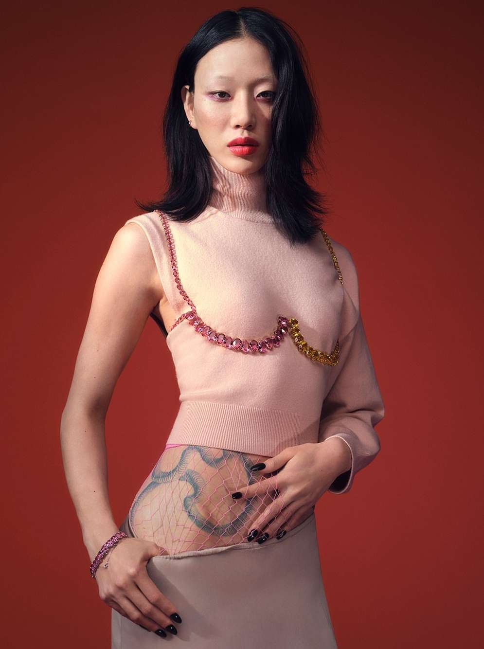 Asian woman poses in a pale pink top with gemstone detail, a pale skirt, and a sheer hip covering on a red background in a studio shot.