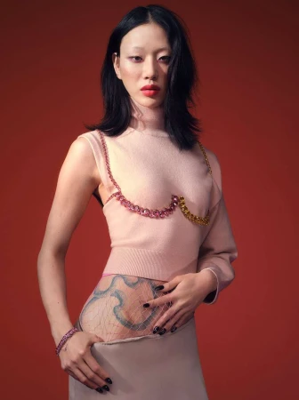 Asian woman poses in a pale pink top with gemstone detail, a pale skirt, and a sheer hip covering on a red background in a studio shot.