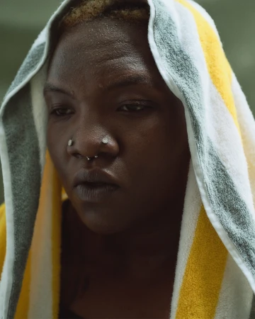 Close-up of a person with dark skin and facial piercings, wearing a striped towel over their head, looking downward in natural light.