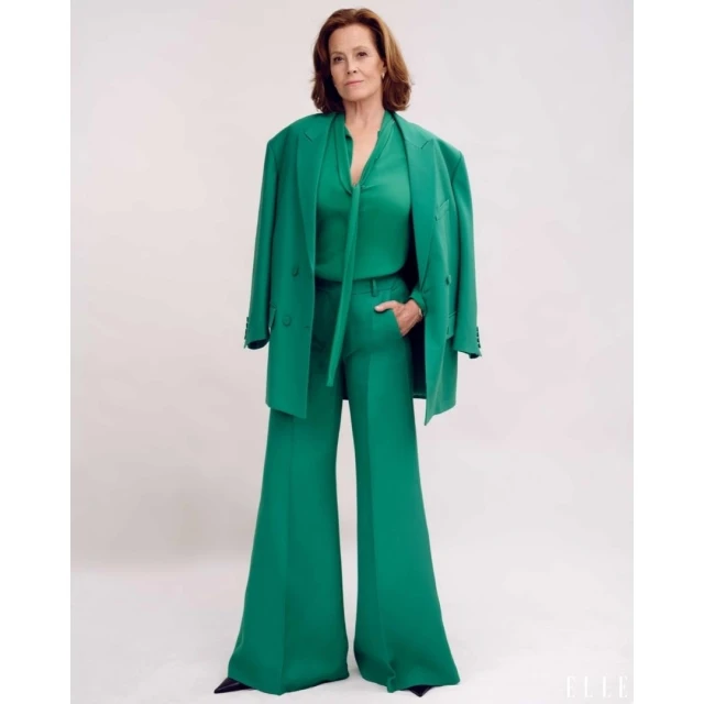 Elegant older woman in a green pantsuit with matching blouse and jacket, standing in a photo studio with a simple, light backdrop.