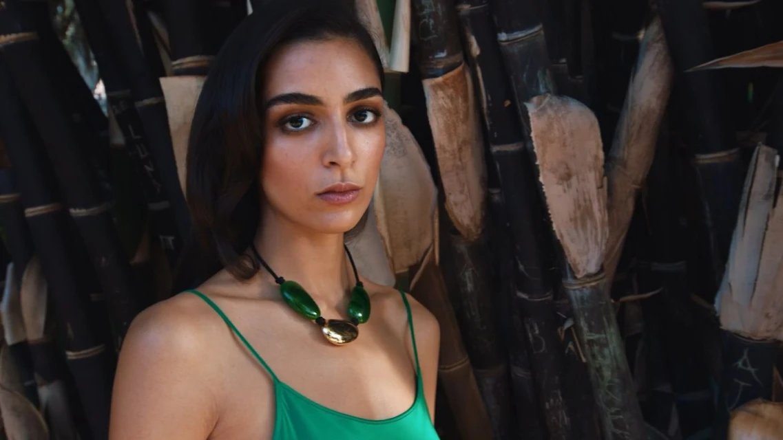 Portrait of a woman with brown hair wearing a green top and a statement necklace, posing against a backdrop of dark bamboo stalks.