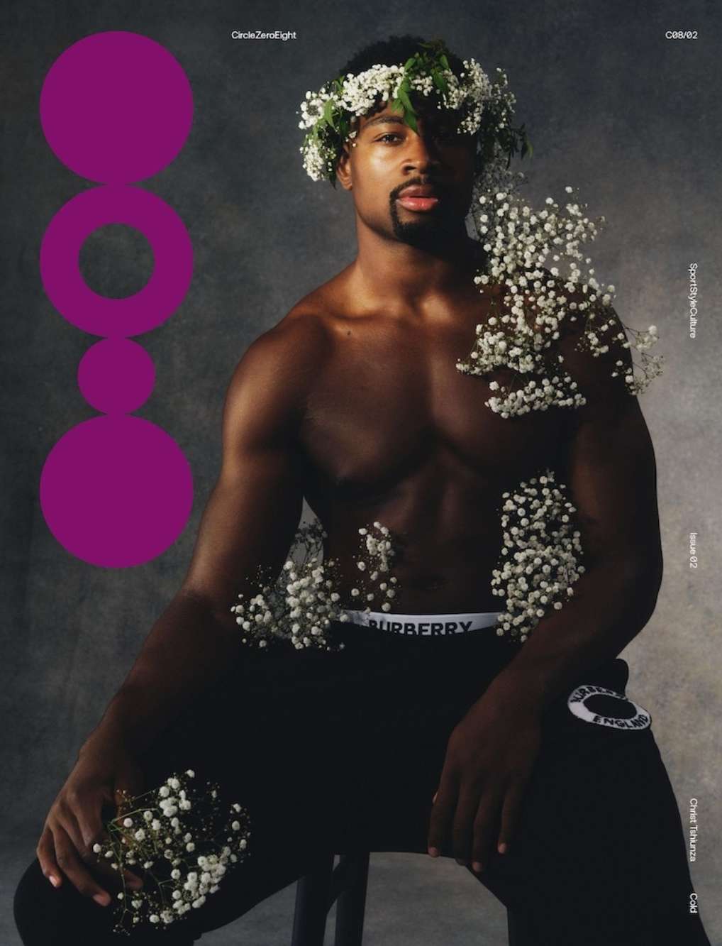 Portrait of a Black man in athletic wear with a floral wreath, seated against a smoky grey backdrop, accented by scattered white flowers.