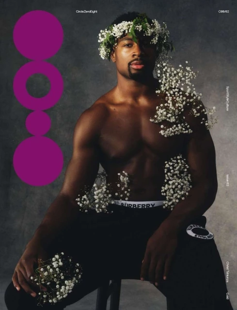 Portrait of a Black man in athletic wear with a floral wreath, seated against a smoky grey backdrop, accented by scattered white flowers.