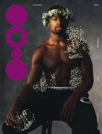 Portrait of a Black man in athletic wear with a floral wreath, seated against a smoky grey backdrop, accented by scattered white flowers.