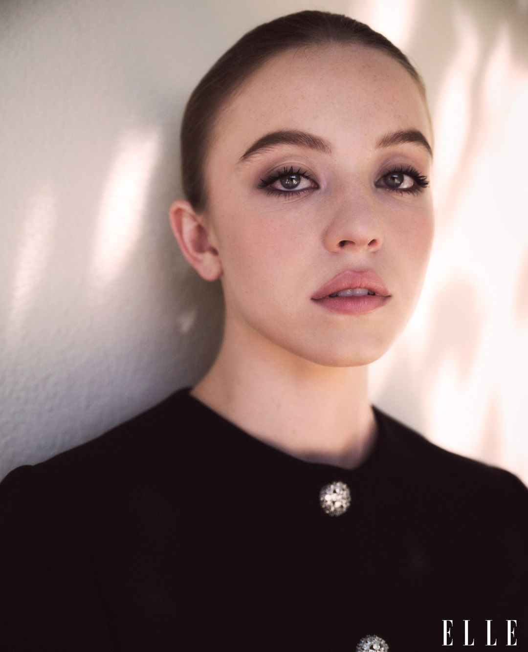 Close-up portrait of a young adult woman with dark eye makeup and a black top with decorative buttons, set against a neutral wall.
