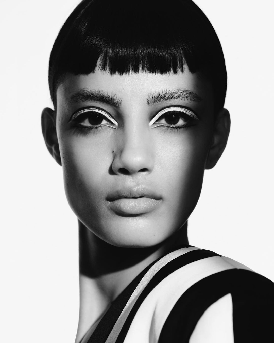 Striking black-and-white close-up of a woman with short dark hair and white eyeliner, wearing a striped top against a light background.