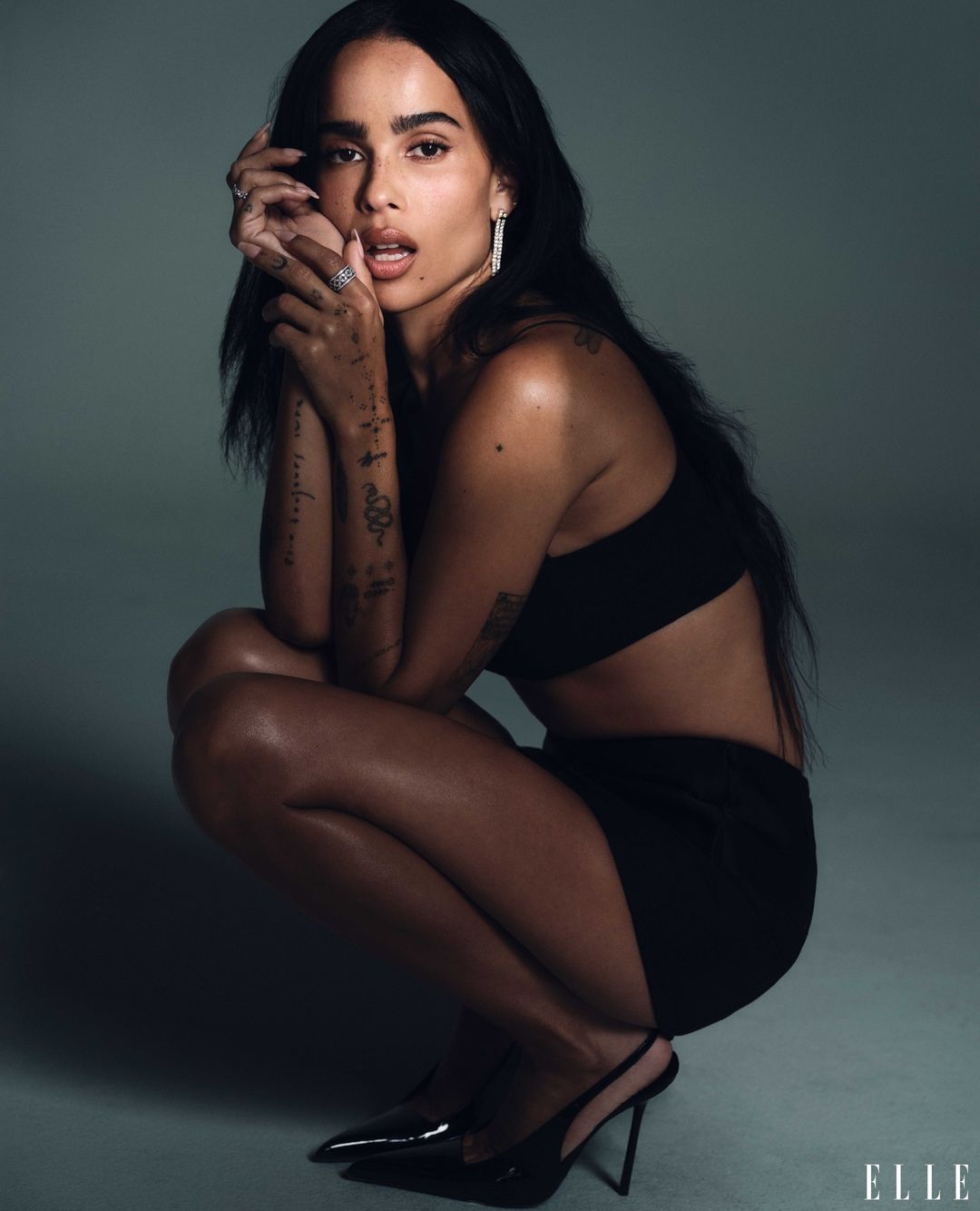 Zoë Kravitz poses in a photo studio, squatting in a black crop top, a black mini skirt, and black high heels against a neutral gray backdrop.