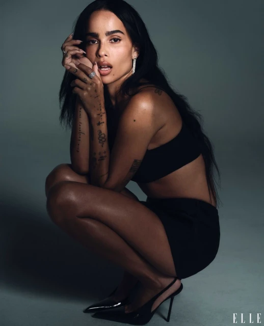 Zoë Kravitz poses in a photo studio, squatting in a black crop top, a black mini skirt, and black high heels against a neutral gray backdrop.