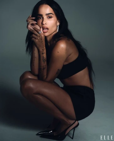 Zoë Kravitz poses in a photo studio, squatting in a black crop top, a black mini skirt, and black high heels against a neutral gray backdrop.
