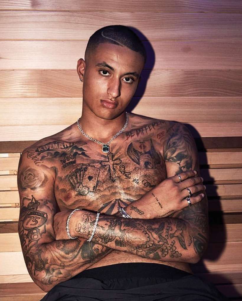 Shirtless tattooed man posing in a sauna with wood-paneled walls, wearing a necklace and bracelets, looking directly at the camera.