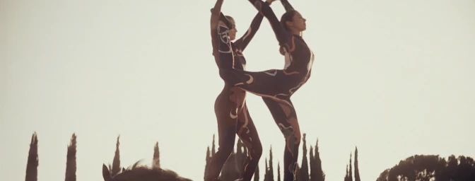 Two women in matching printed bodysuits performing an acrobatic pose outdoors against a silhouetted treeline at sunset.