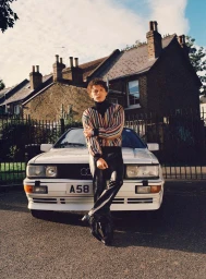 Tom Holland leaning against a white vintage car wearing a striped sweater and dark trousers