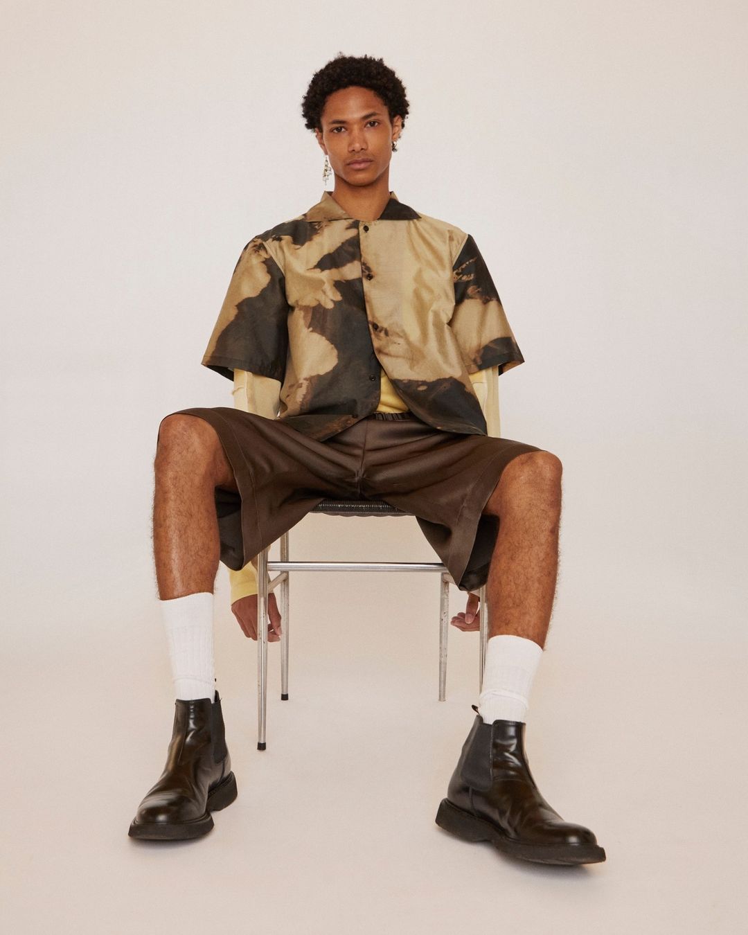 Young man wearing a short-sleeve patterned shirt, brown shorts, and black boots seated on a metal chair in a studio.
