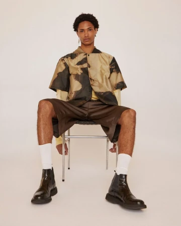Young man wearing a short-sleeve patterned shirt, brown shorts, and black boots seated on a metal chair in a studio.
