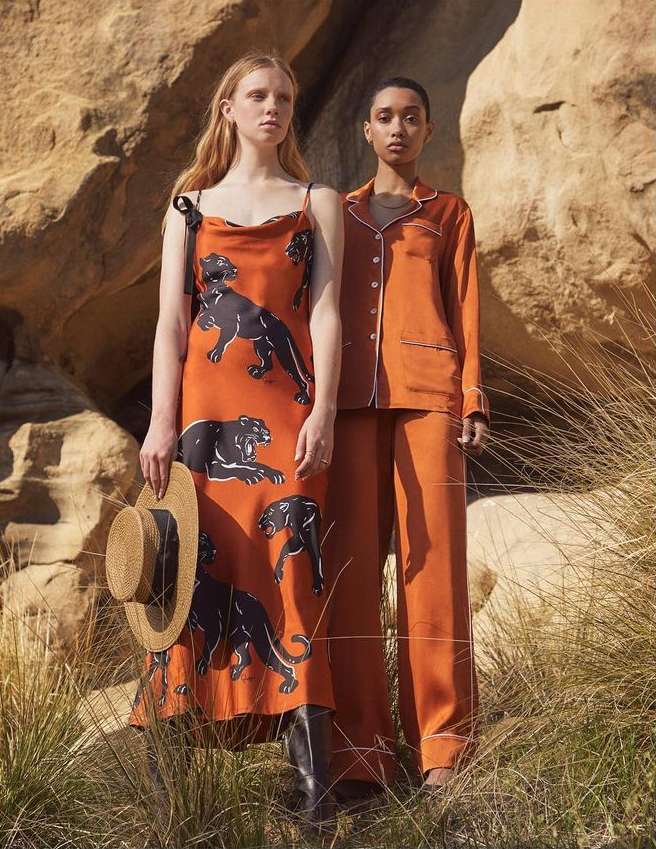 Two women model orange dresses with panther prints in a desert setting. One holds a straw hat and wears boots; the other wears an orange suit.