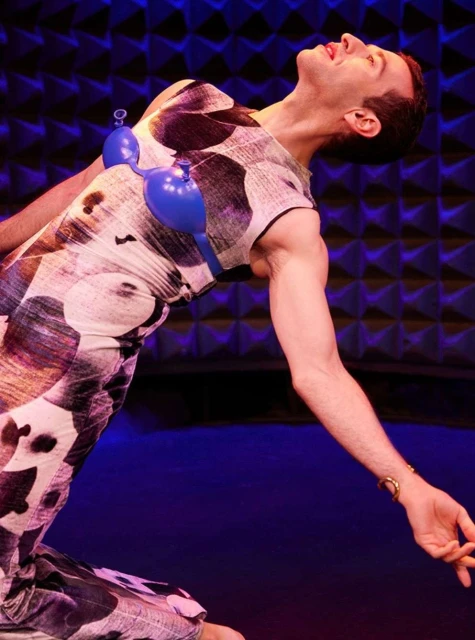 A person in patterned dress strikes a theatrical pose on stage in a blue-lit performance, with blue balloons on their chest.