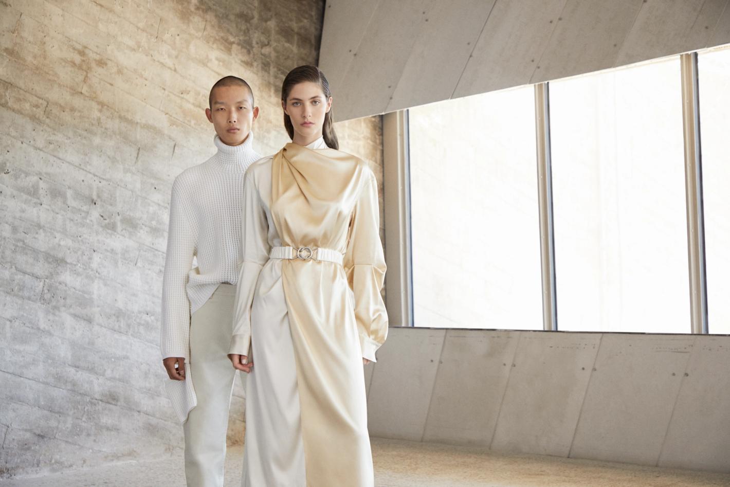 Fashion editorial shot of a man in a white turtleneck and a woman in a cream-colored dress in front of a concrete wall and window.