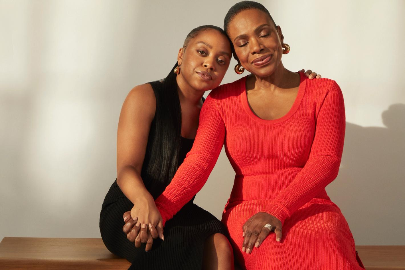 Portrait of a smiling adult woman with her arm around a mature adult woman sitting on a bench in front of a white background.
