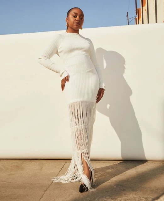 Woman in a long-sleeved white dress with fringe detail poses in front of a white wall, showcasing a blend of elegance and modern style.