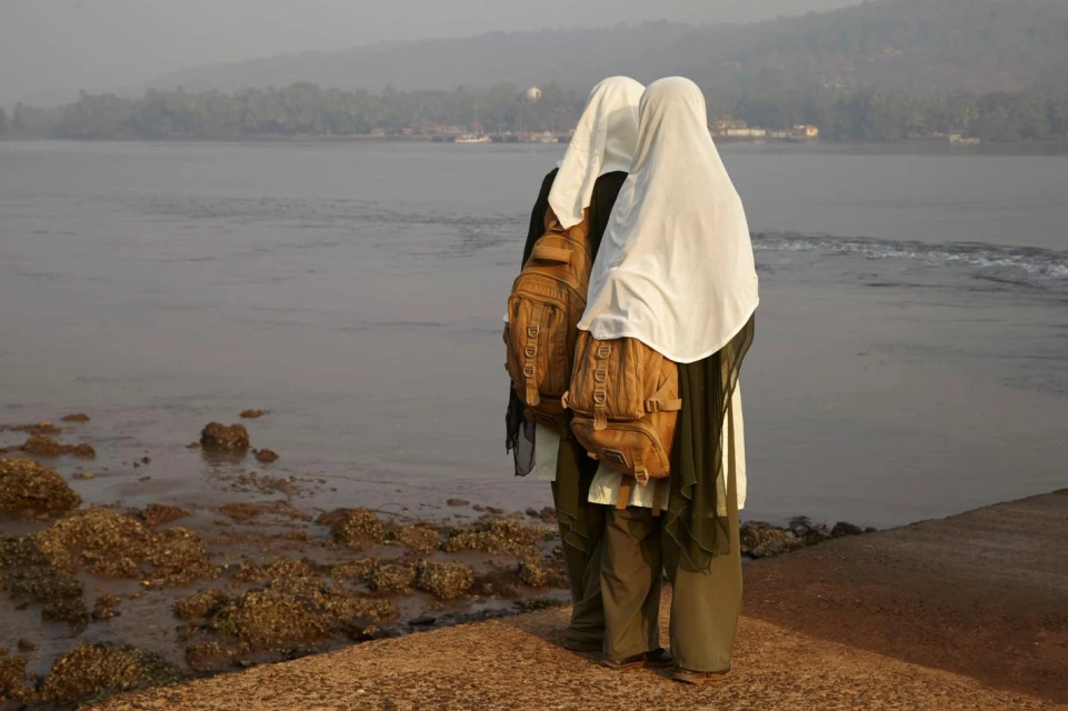 Two veiled women with backpacks stand near a body of water with a shoreline of rocks and trees under an overcast sky.