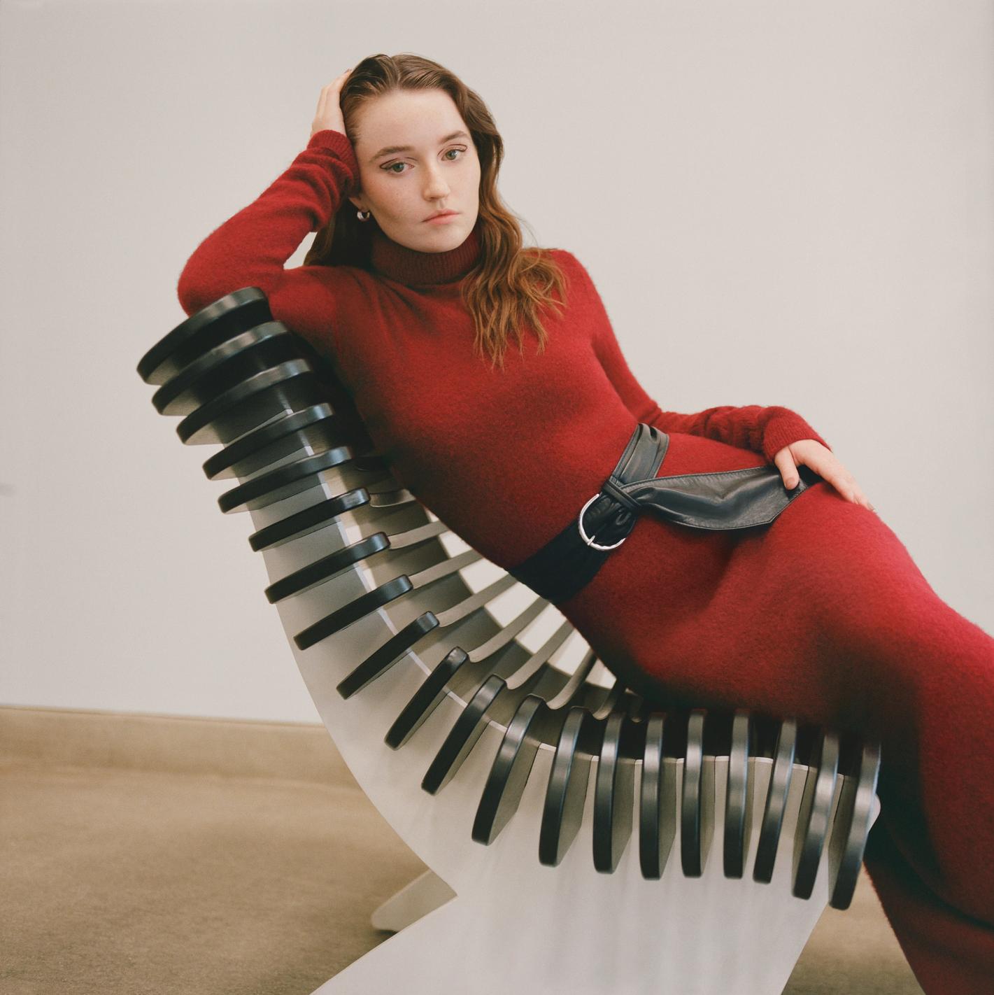 A woman in a red dress reclining on a modern chair with a white background. She has her hand on her head and a belt on the dress.